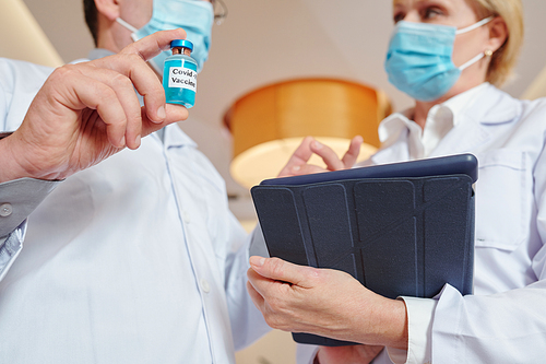 Close-up image of researher with vial and tablet computer discussing process of new vaccine testing