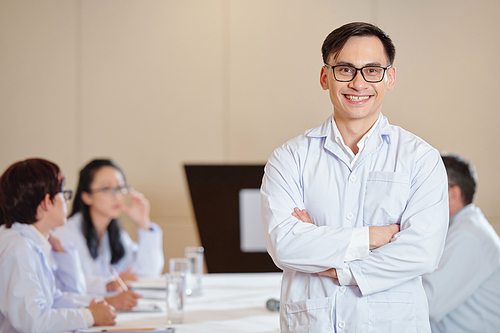 Portrait of happy smiling young scientist in labcoat standing at meeting table with arms folderd and looking at camera