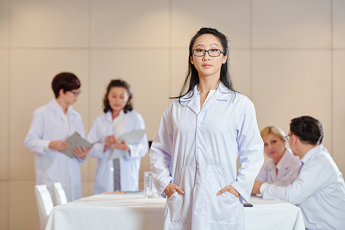 Portrait of young serious Chinese researcher in white labcoat standing in conference room, her colleagues talking in background