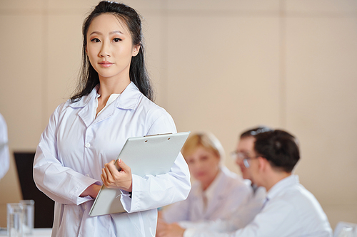 Serious young female Chinese scientist with clipboard standing in conference room and looking at camera