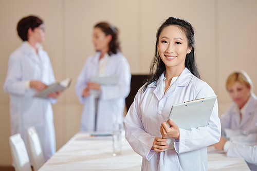 Portrait of lovely attractive young Chinese medical researcher standing clipboard and smiling at camera, her colleagues talking in background