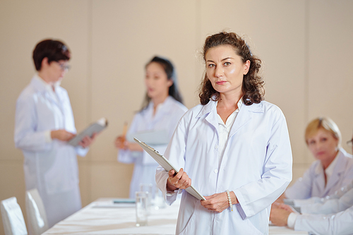 Portrait of serious mature female researcher holding clipboard and looking at camera when standing in conference room