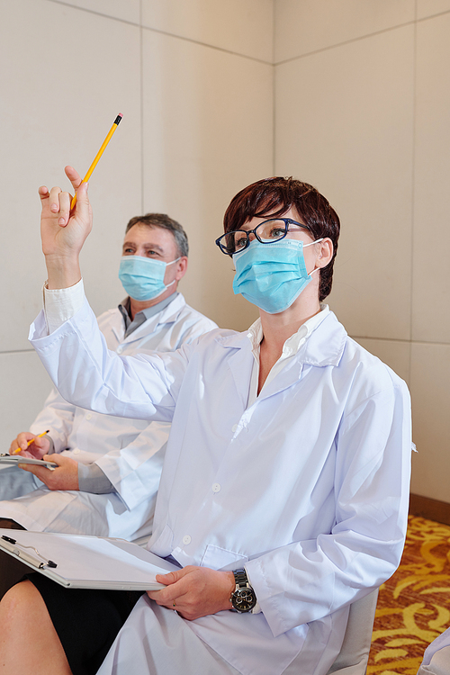 Young female scientist in medical mask attending conference and raising hand to ask speaker a question