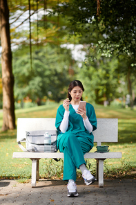 Medical nurse sitting on bench in park, eating snacks and reading posts on social media