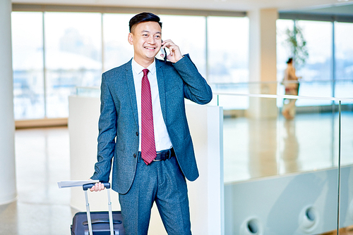 Portrait of modern Asian businessman speaking by phone and smiling happily while waiting in airport holding suitcase