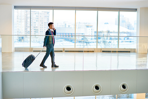 Side view portrait of modern Asian businessman carrying suitcase and speaking by phone while crossing airport waiting lounge, copy space