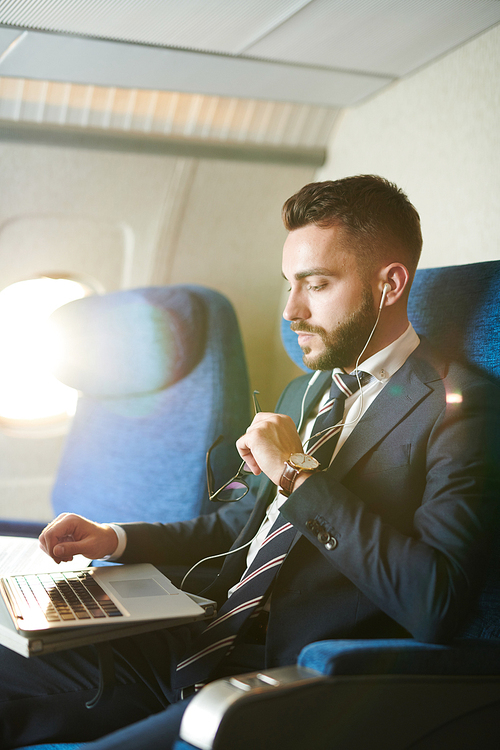 Side view portrait of handsome bearded businessman using laptop wearing earphones while enjoying first class flight in plane, copy space