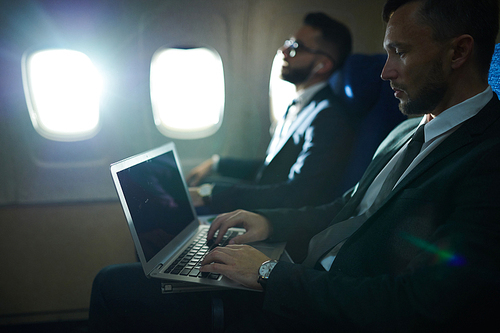 Side view portrait of two business people in first class of dimly lit plane, focus on man using laptop in front, copy space