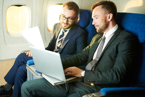 Portrait of two successful businessmen using laptop and working while enjoying first class flight in plane copy space