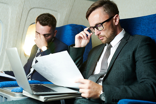 Portrait of two tired business people working and using laptop in plane during first class flight, copy space