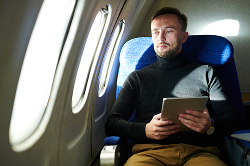 Portrait of mature handsome man sitting in first class and looking at window pensively while holding digital tablet, copy space