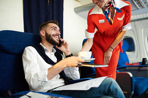 Portrait of smiling flight attendant serving coffee to handsome businessman speaking by phone in first class, copy space