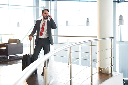 Full length portrait of handsome bearded businessman with suitcase talking to his colleague on smartphone while standing at airport and waiting for departure
