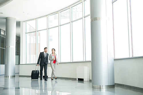 Full length portrait of successful couple, man and woman, walking across hall of modern airport carrying suitcase, copy space background