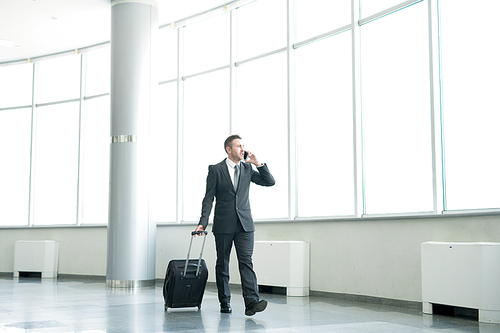 Full length portrait of successful businessman walking across hall of modern airport carrying suitcase and speaking by phone, copy space background