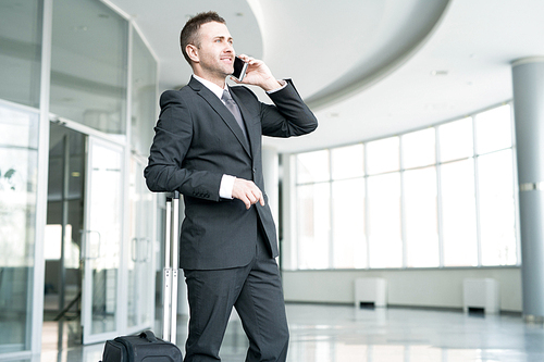 Portrait of successful businessman standing in hall of modern airport and speaking by phone smiling cheerfully