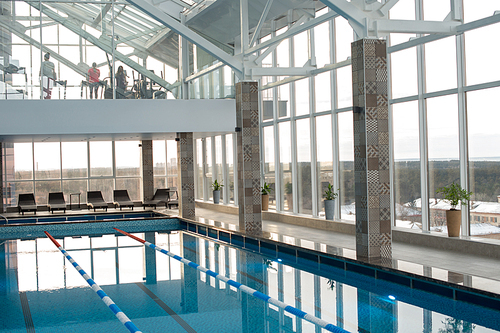 Interior of spacious modern wellness center with glass walls: swimming pool and deck chairs on first floor, fitness hall with visitors on second one