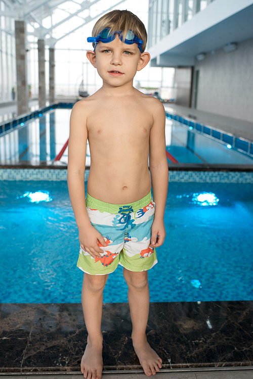 Confident cute boy in swimming shorts standing on edge of swimming pool and  in indoor pool with clean water