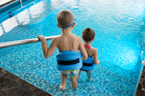 Back view of unrecognizable little children wearing swimming belts going down to pool while having lesson with their coach