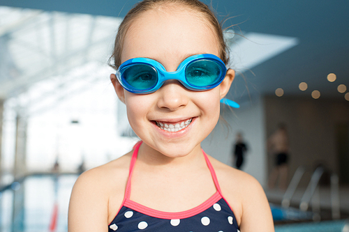 Head and shoulders portrait of cheerful little girl wearing swimsuit and goggles  with toothy smile while spending weekend in swimming pool, blurred background