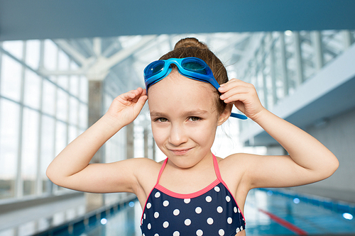 Cute little girl wearing swimsuit and goggles posing for photography while standing on edge of swimming pool, head and shoulders portrait shot