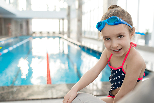 Pretty little girl wearing swimsuit and goggles sitting on deck chair and posing for photography while taking break from swimming in spacious pool, portrait shot