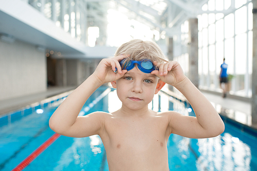 Calm cute boy with blond hair training in swimming pool: he putting on goggles and 