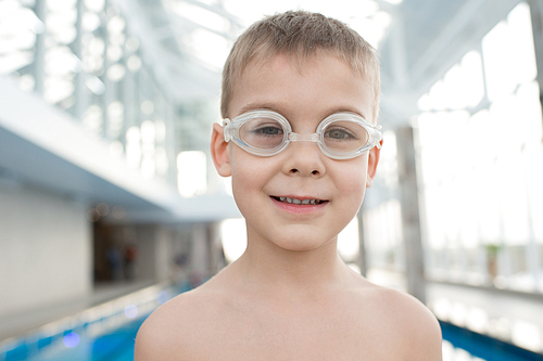 Portrait of cheerful cute naked kid in transparent swimming goggles smiling at camera and standing in indoor pool