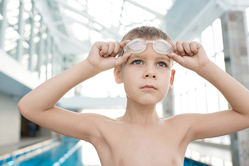Serious determined male kid looking into distance and preparing for training in swimming pool, he adjusting goggles