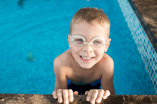 Portrait shot of cute little boy in swimming goggles  with toothy smile while holding edge of pool