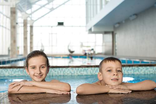 Cheerful positive children leaning on edge of swimming pool and  optimistically enjoying leisure in swimming