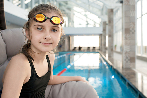 Portrait of serious confident pretty girl with wet hair wearing goggles and athletic swimwear sitting in comfortable armchair by spacious swimming pool