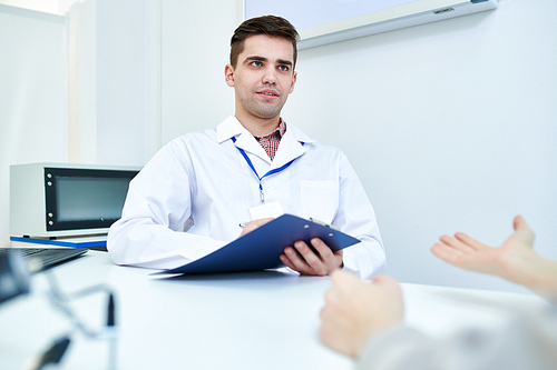 Portrait of young doctor holding clipboard  listening to patients symptoms while sitting at desk in office, copy space