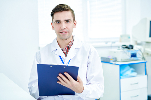 Waist up portrait of young doctor holding clipboard and  posing in modern office, copy space