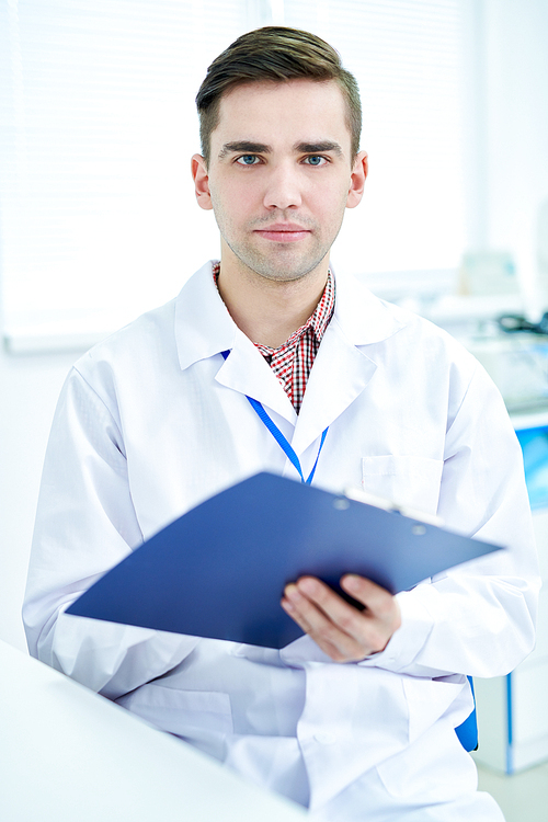 Waist up portrait of confident young doctor holding clipboard and  posing in modern office