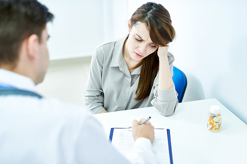 Portrait of frowning young woman listening to doctor while sitting at desk in clinic, copy space