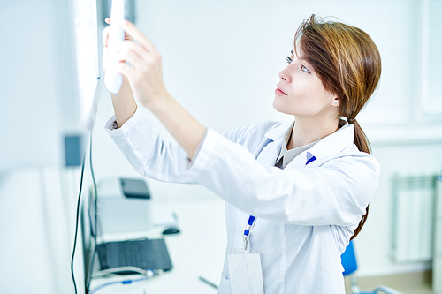 Side view of female medic standing in white coat and looking at X-ray image holding it against lightbox.