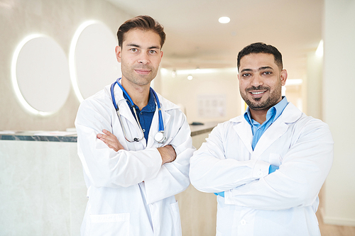 Waist up portrait of two doctors, one of them Middle-Eastern,  and smiling while posing in modern clinic, copy space