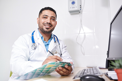 Portrait of mature Middle-Eastern doctor wearing lab coat  while posing with clipboard at desk in office, copy space