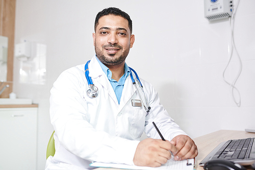Portrait of mature Middle-Eastern doctor wearing lab coat  while posing at desk in office, copy space