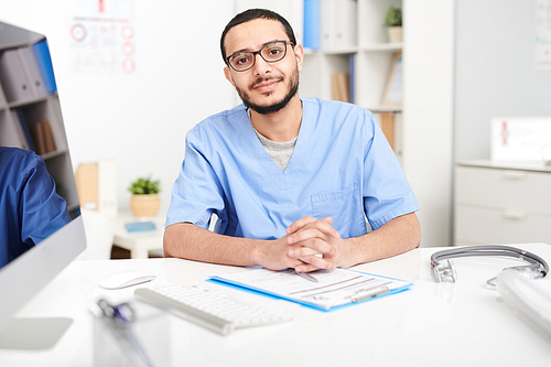 Portrait of young Middle-Eastern doctor wearing glasses sitting at desk in office smiling and 