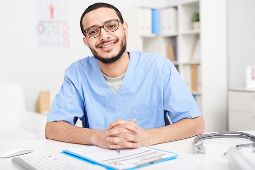 Portrait of  smiling young Middle-Eastern doctor wearing glasses sitting at desk in office and 