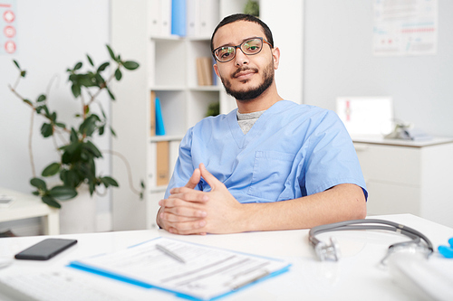 Portrait of confident  young Middle-Eastern doctor wearing glasses sitting at desk in office and 