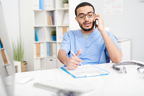 Portrait of young Middle-Eastern doctor wearing glasses sitting at desk in office and speaking by smartphone, filling in patientts forms