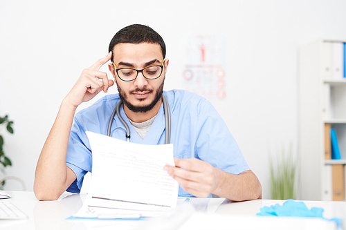 Portrait of  Middle-Eastern doctor wearing glasses sitting at desk in office and reading patients reports