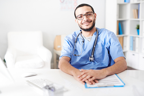 Portrait of Middle-Eastern friendly doctor wearing glasses sitting at desk in office and  smiling happily, copy space