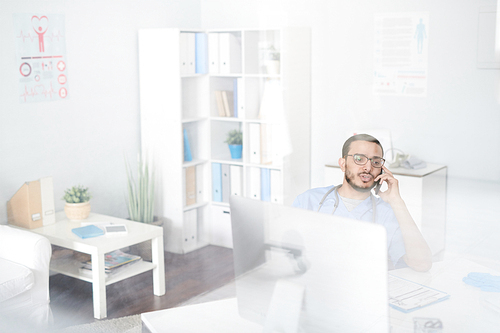 Portrait of young Middle-Eastern doctor wearing glasses sitting at desk in office and speaking by smartphone, shot from behind glass wall, copy space