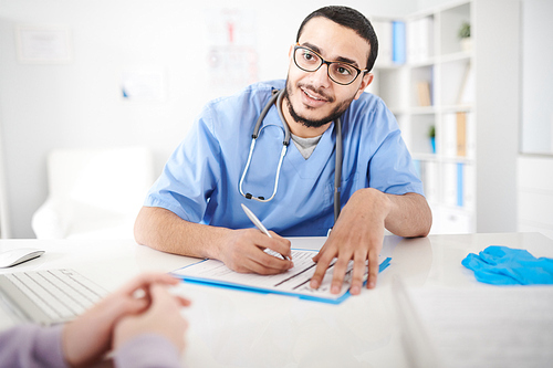 Portrait of young Middle-Eastern doctor wearing glasses sitting at desk in office and listening intently to unrecognizable patient while making notes on clipboard