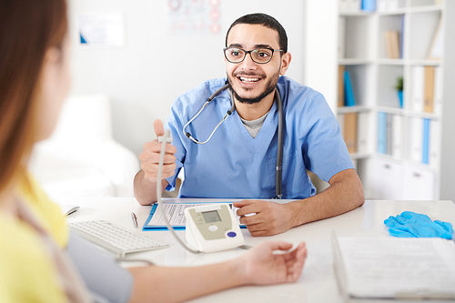 Portrait of young Middle-Eastern doctor wearing glasses sitting at desk in office and measuring blood pressure of female patient