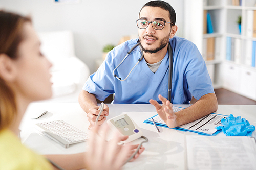 Portrait of young Middle-Eastern doctor wearing glasses talking to female patient sitting at desk in office of modern clinic